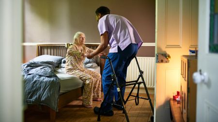 Young male care worker helping elderly woman off bed in a nursing home