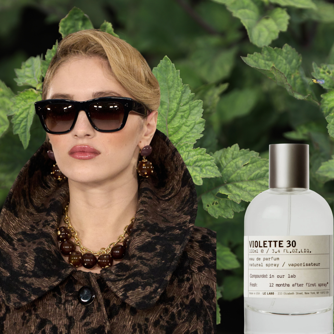 A blonde woman wearing oversized sunglasses, a brown coat, and brown jewelry. She is in front of a background of patchouli leaves and is next to three perfume bottles from Le Labo, Dolce and Gabbana, and Krigler