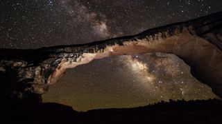 a large rock structure bridges across the image in front of the Milky Way.