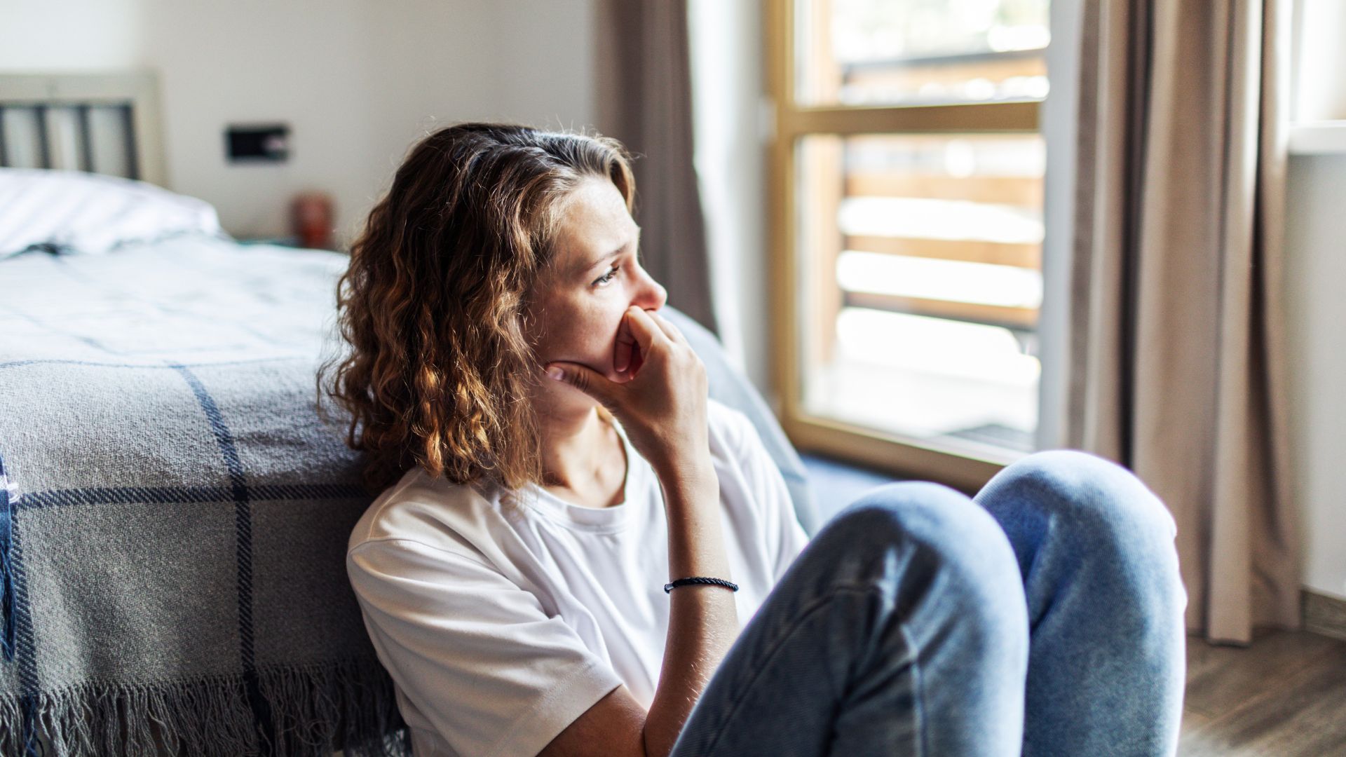 A woman sits on the floor next to her bed looking upset and emotional.