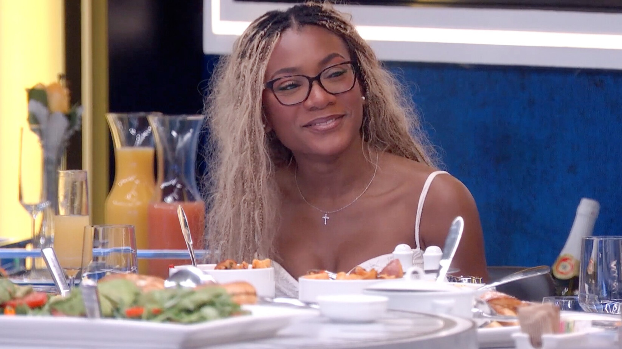 Taylor sitting at the time travel table enjoying a lavish meal. Her hair is down and she's smiling. 