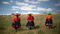 three women wearing red shirts and hats walk through a grassy field towards a mountain
