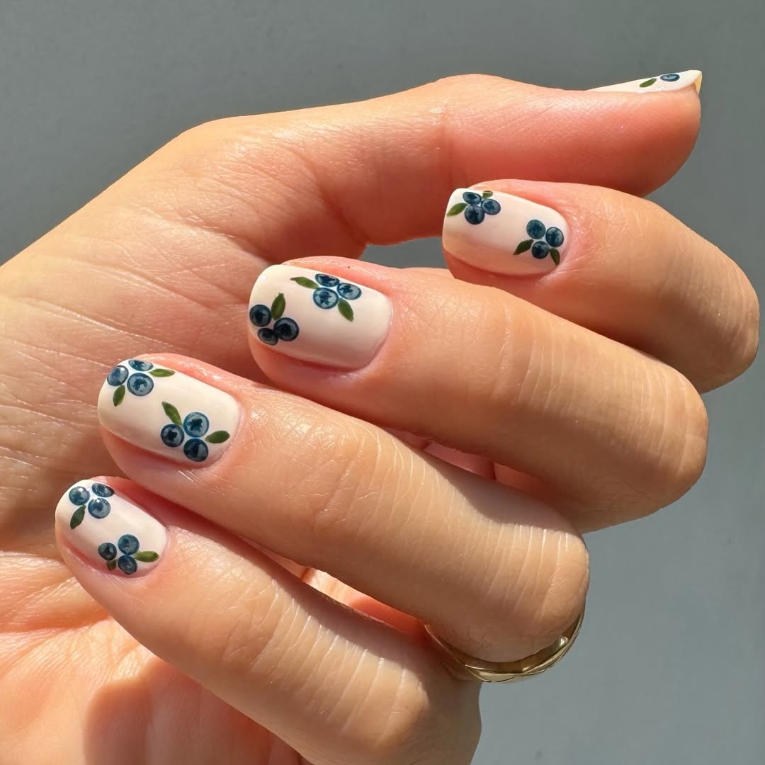 A hand with a nude-painted nail base covered in tiny hand-drawn blueberries.