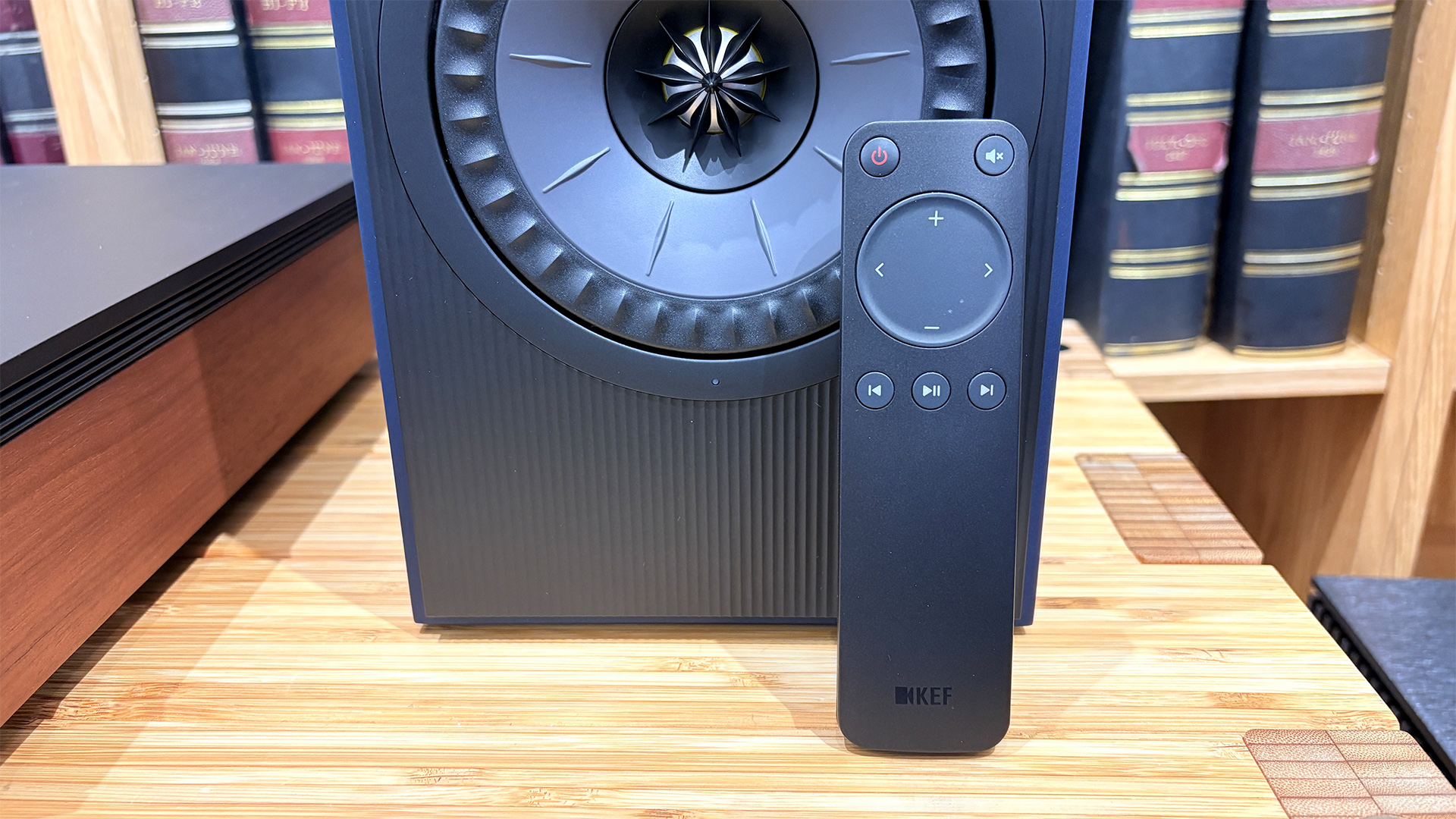 KEF Coda W active speakers on wooden hi-fi rack in front of bookcase with remote control