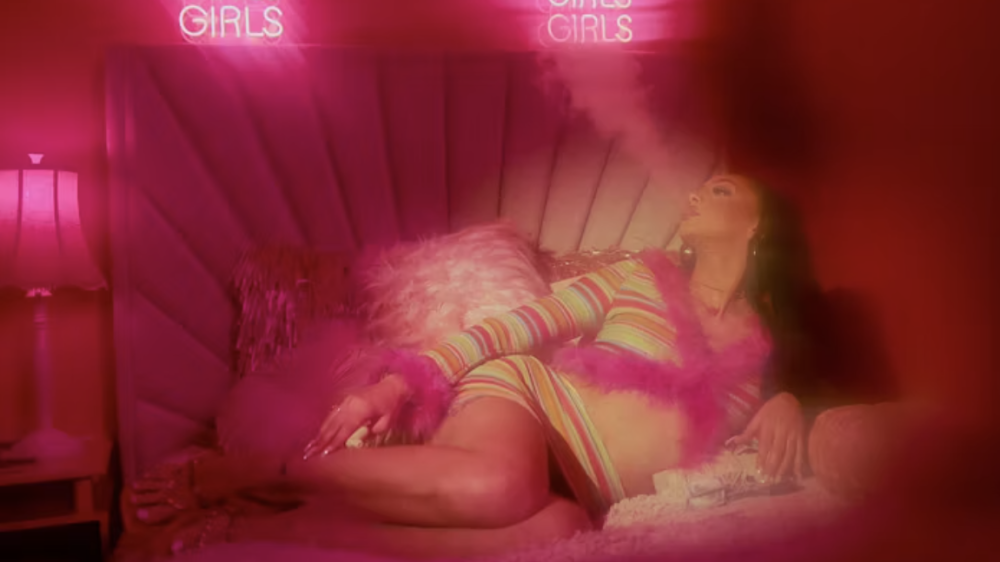A woman smoking on a bed beneath pink neon signs in Magic City: An American Fantasy