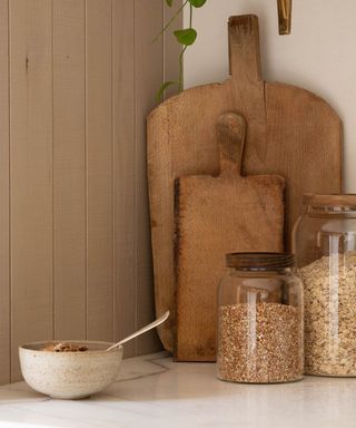 A set of three large glass storage jars with wooden lids in front of two wooden cutting boards and next to an stonewarebowl of cereal
