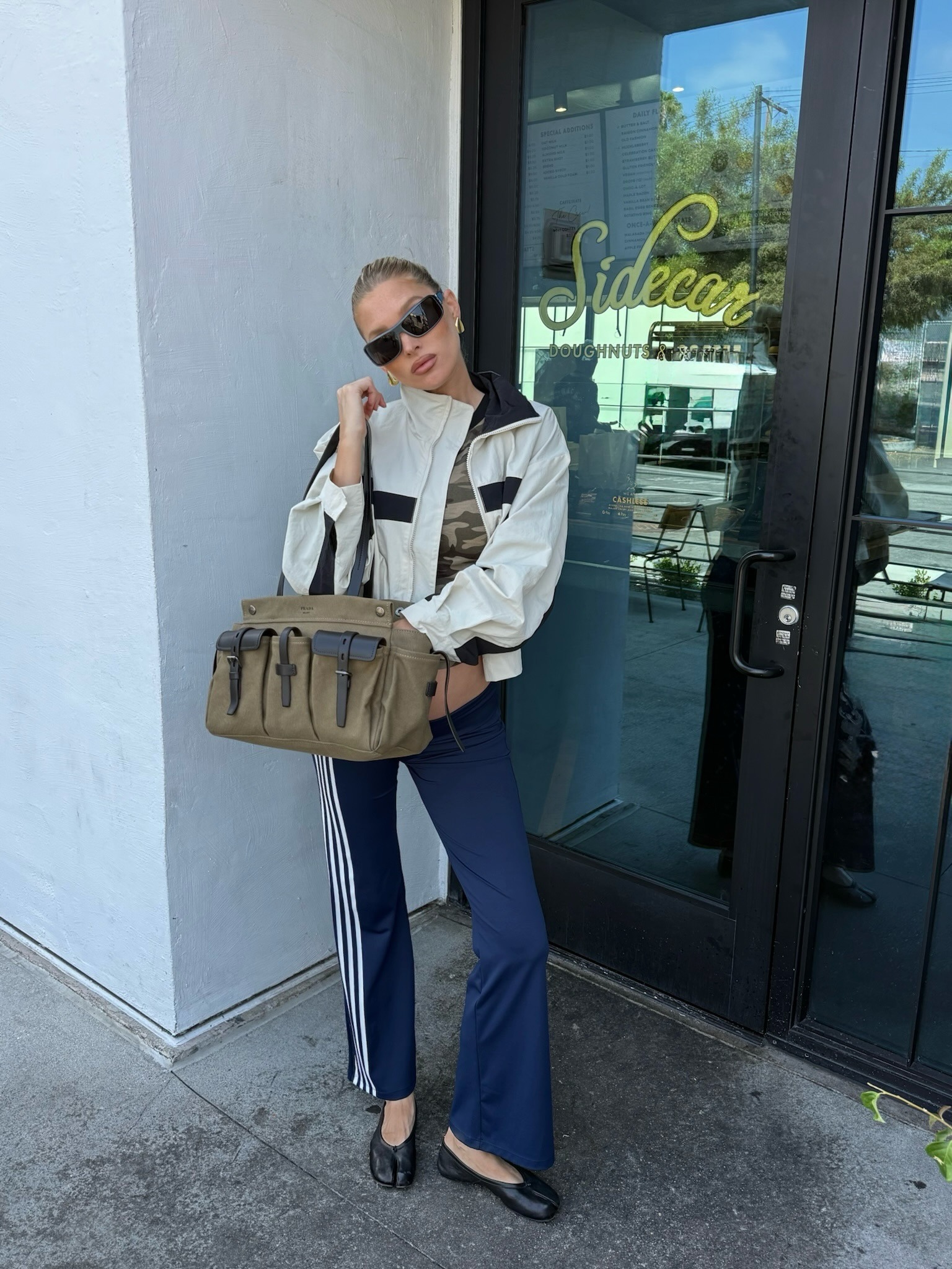 Elsa Hosk wearing a windbreaker, a camo T-shirt, Adidas track pants, Maison Margiela Tabi ballet flats, and a Prada Route bag.