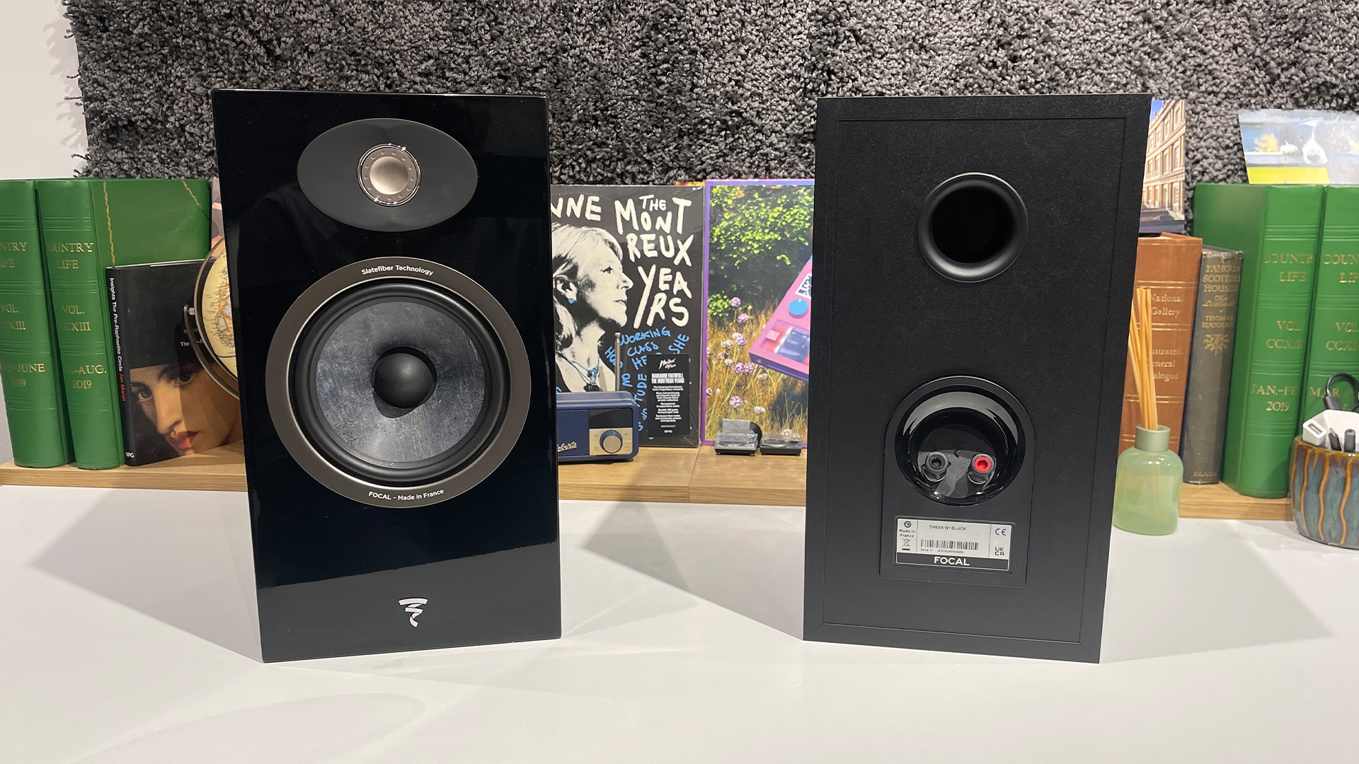 Focal Theva No.1 standmount speakers on white wooden table in front of busy bookshelf, one of the cabinets is rear-facing