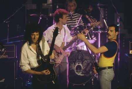 Queen perform live in 1986, with Freddie Mercury wearing a yellow basketball vest over a blue T-shirt, Brian May wearing an open collared white shirt, and John Deacon playing a prototype Warwick Buzzard bass, which was designed by John Entwistle and is up for auction in December 2025.