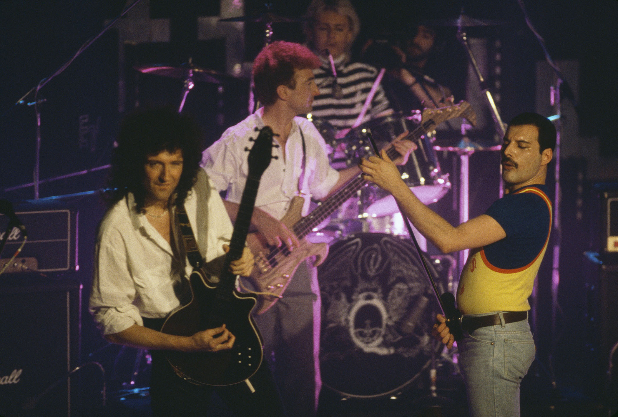 Queen perform live in 1986, with Freddie Mercury wearing a yellow basketball vest over a blue T-shirt, Brian May wearing an open collared white shirt, and John Deacon playing a prototype Warwick Buzzard bass, which was designed by John Entwistle and is up for auction in December 2025.