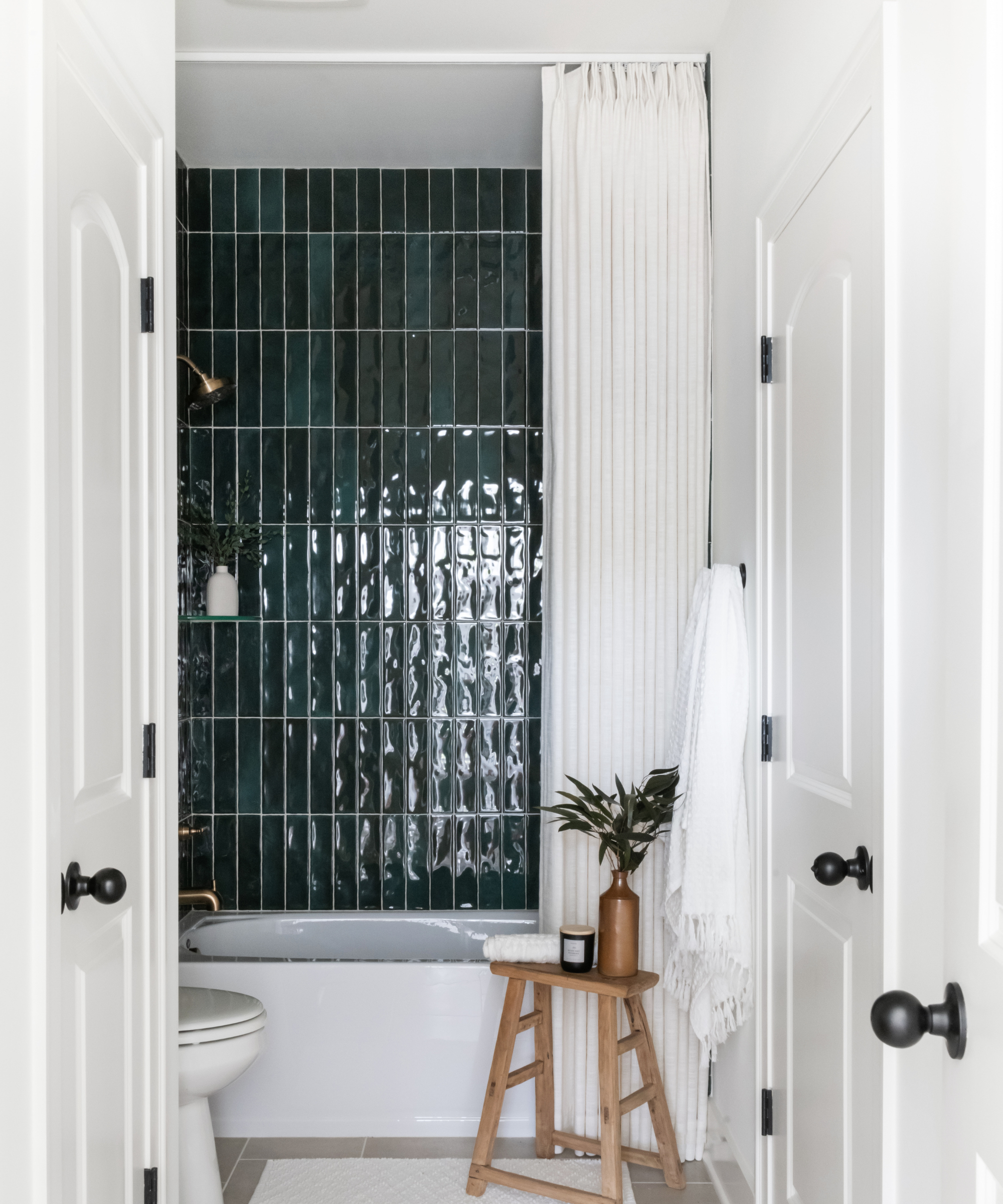 a bathroom with deep green tiling, a white shower curtain, white tub, white walls, and white doors, and a small wooden stool next to the tub