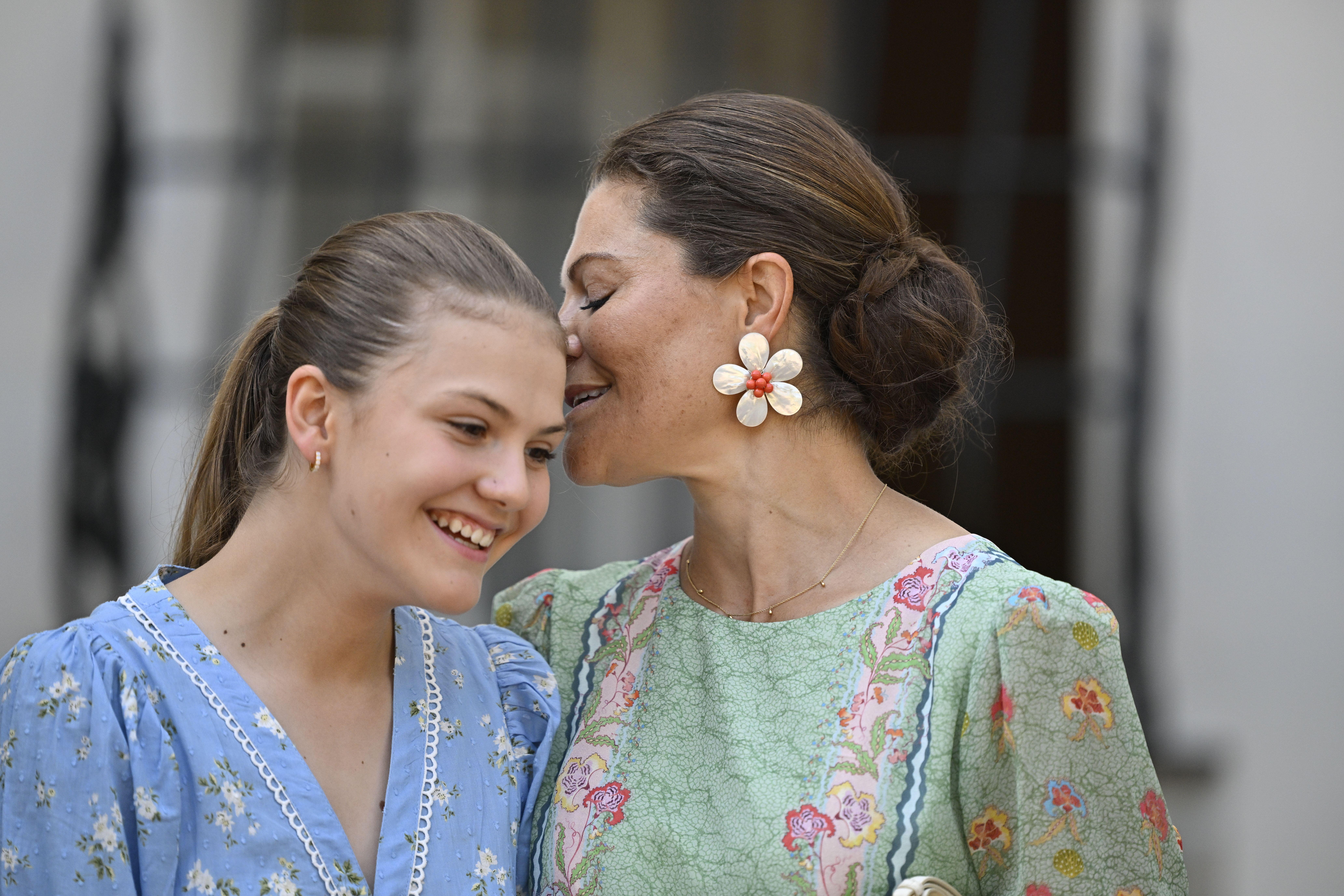 Princess Estelle wearing a blue floral dress leaning over to smile at Crown Princess Victoria wearing a green dress