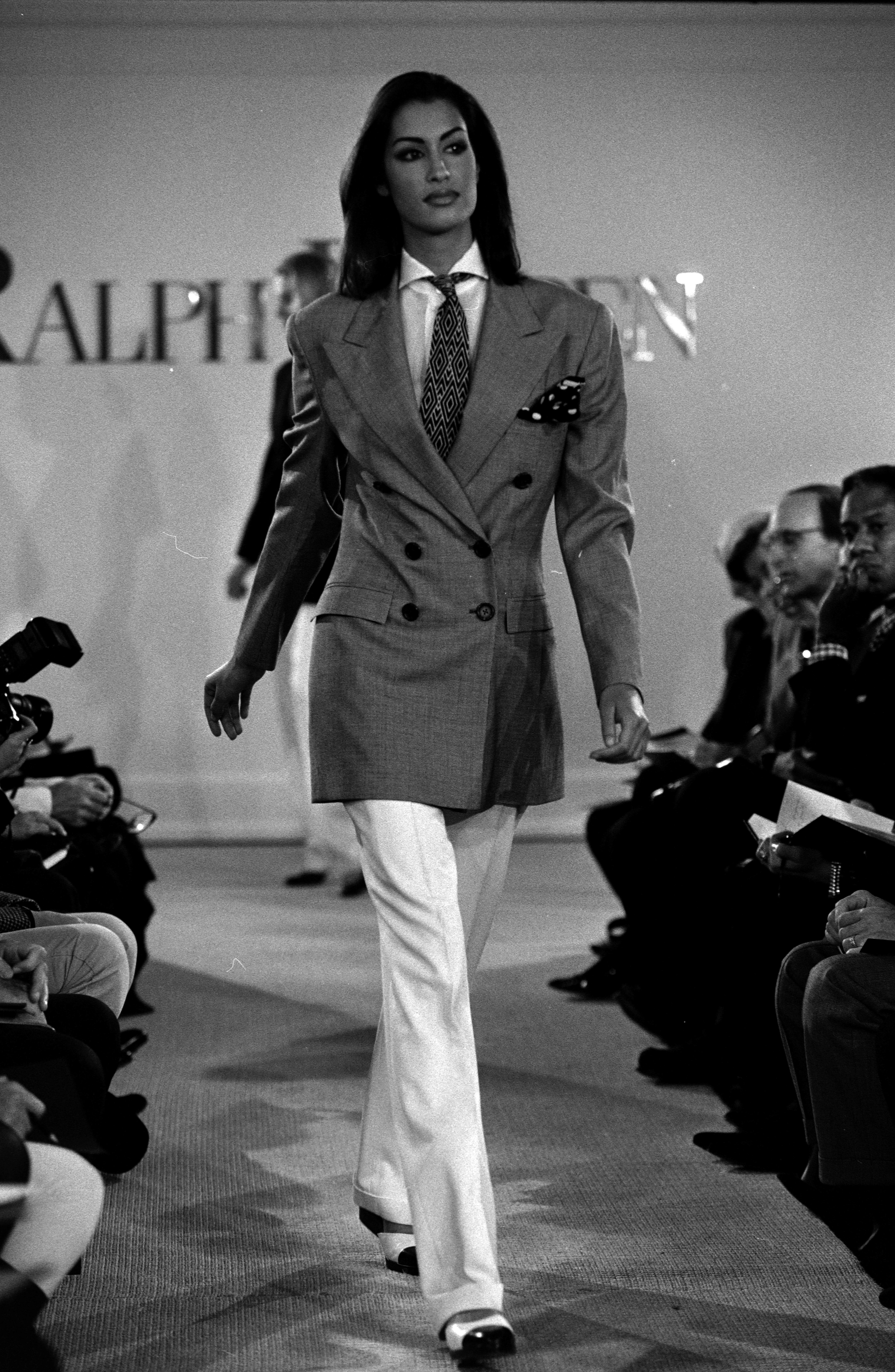 a black-and-white image of a model walking the runway wearing a gray blazer and white pants