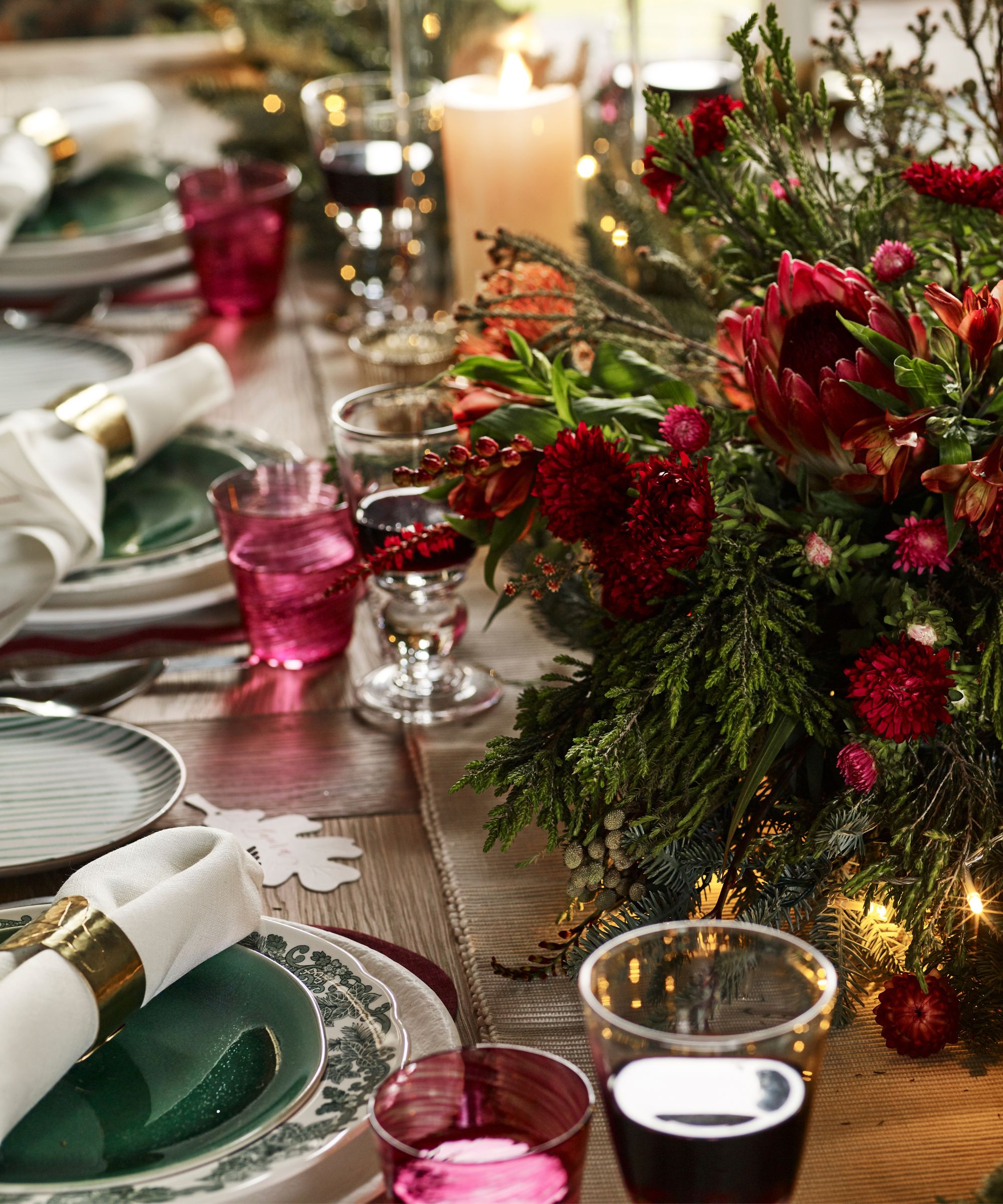 Table with green floral crockery, pink glass tumblers, gold napkin rings, and a pine and red dahlia centerpiece