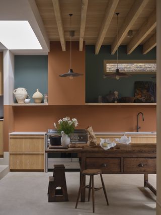 Image of a kitchen with concrete floors and a wooden ceiling. The walls are painted a mix of orange and dark turquoise paint colors. The cabinetry is a light wooden, and there is a rustic wooden kitchen island in the center.