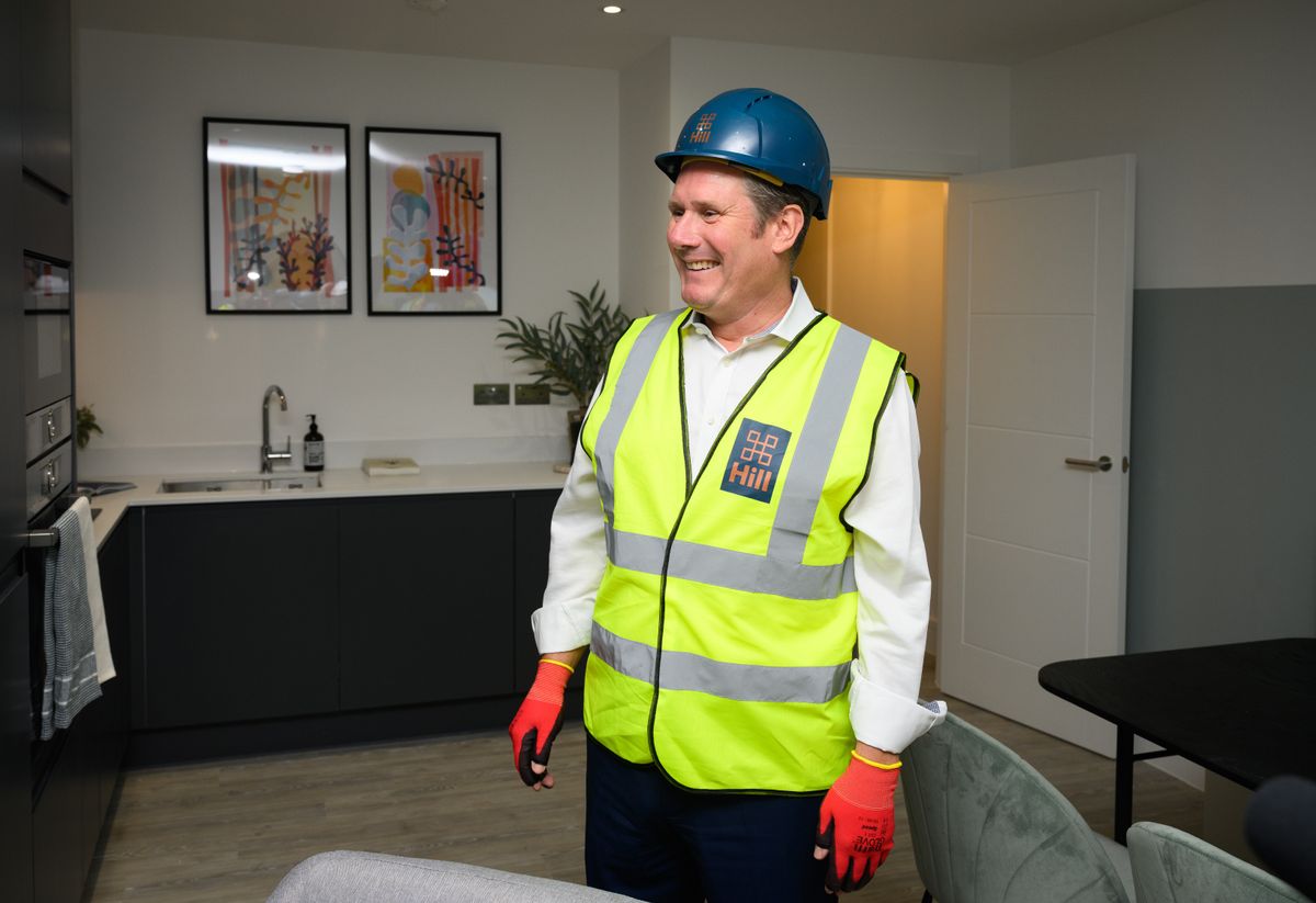 Keir Starmer on a housing development site inside a house&#039;s living room wearing a high-vis jacket and orange gloves and a hard hat