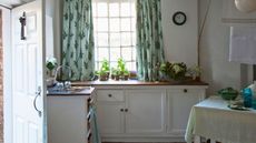 A white vintage style kitchen with wooden countertops, green curtains, white walls, a black clock and dining table covered in a green tablecloth. To the left is an open white front door.