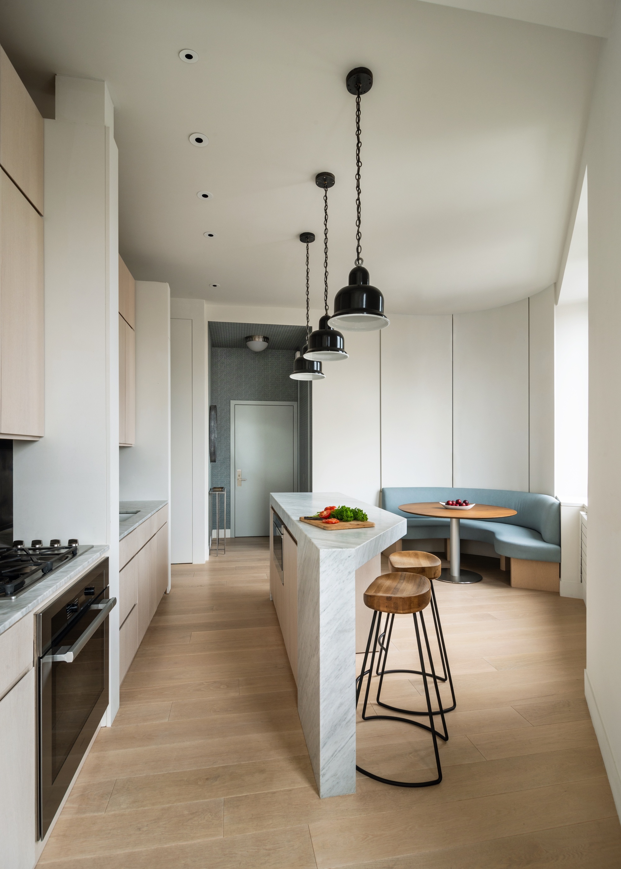 A minimalist kitchen with a tiny angular island, bar stools and banquette seating in the background