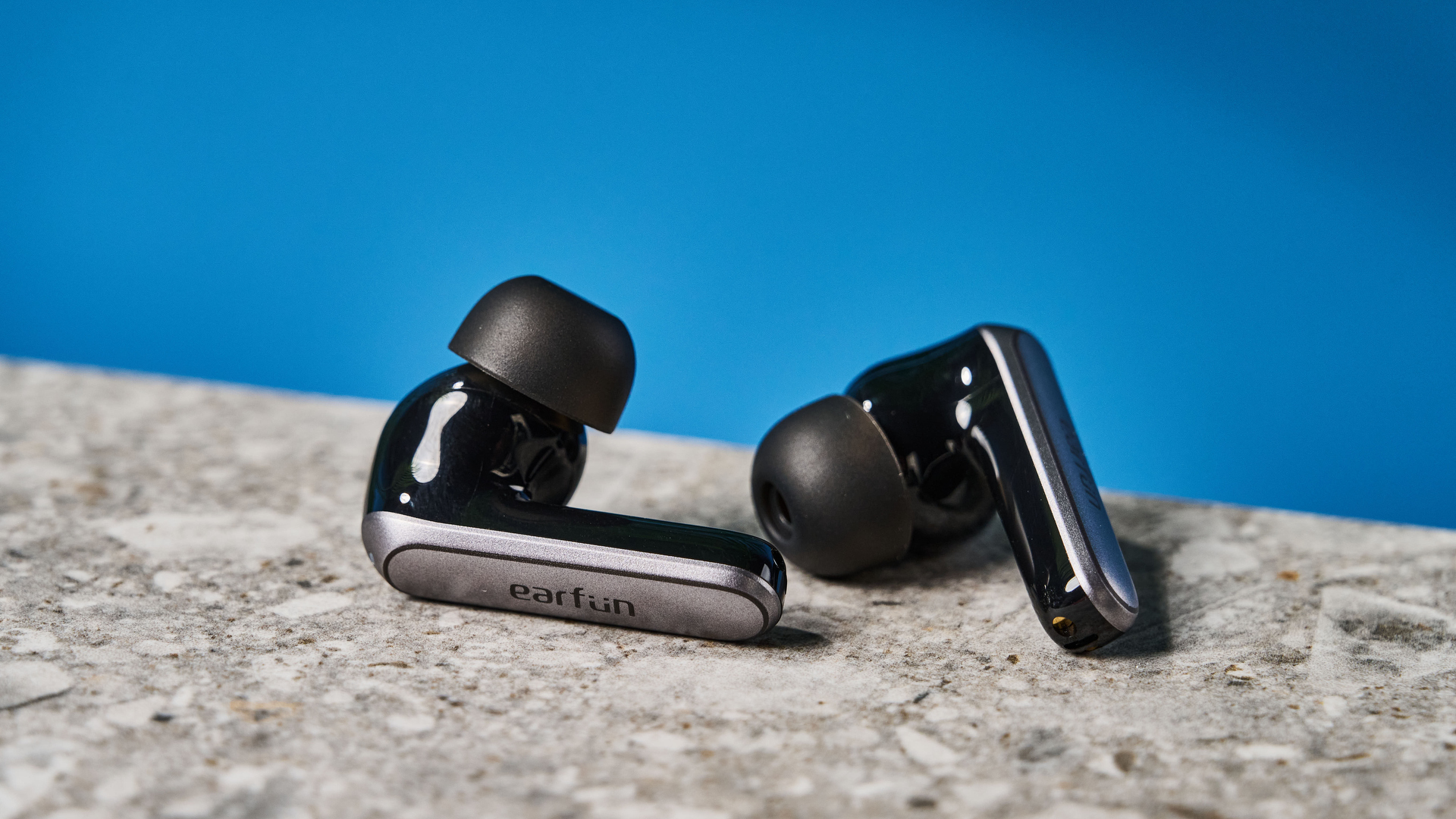 a pair of black and silver earbuds with silicone tips and a black charging case are photographed against a blue background