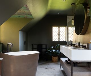 loft conversion bathroom with dark green walls, wallpaper in dormer window, freestanding copper bath set at an angle and large white double sink vanity unit with shelf below