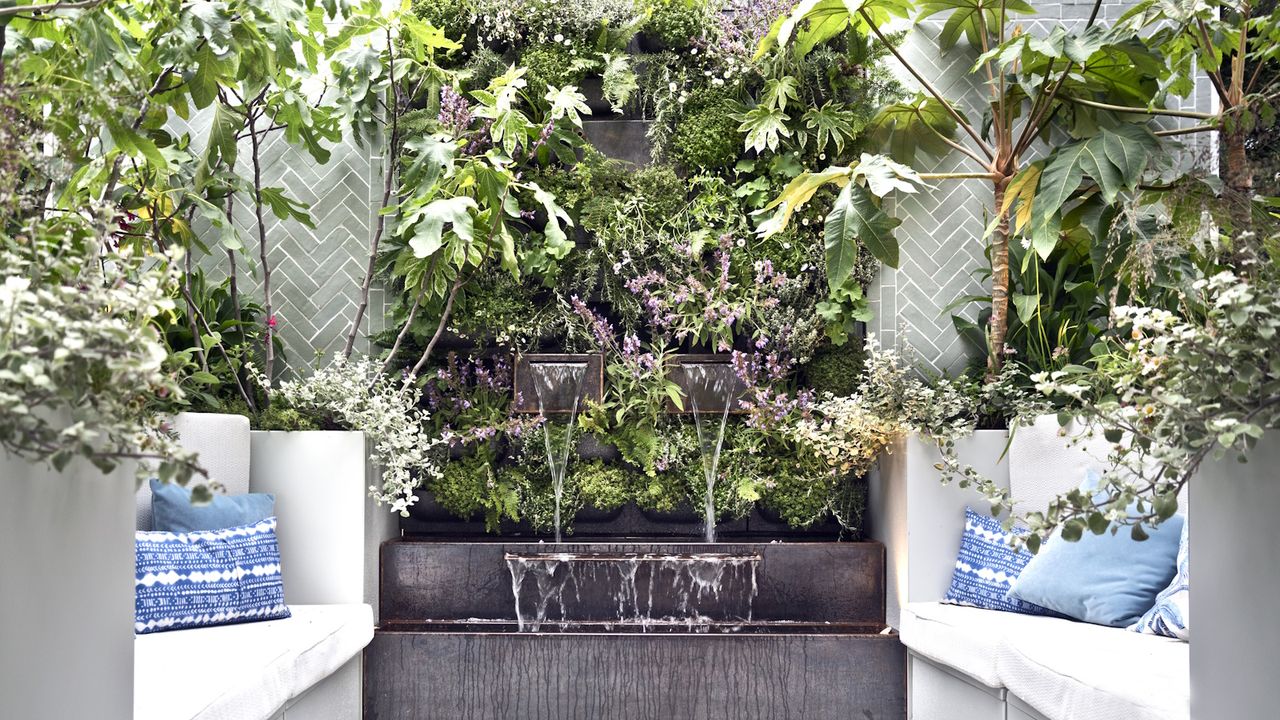 A modern courtyard garden with waterfall, tropical style vertical planting and bench seating either side