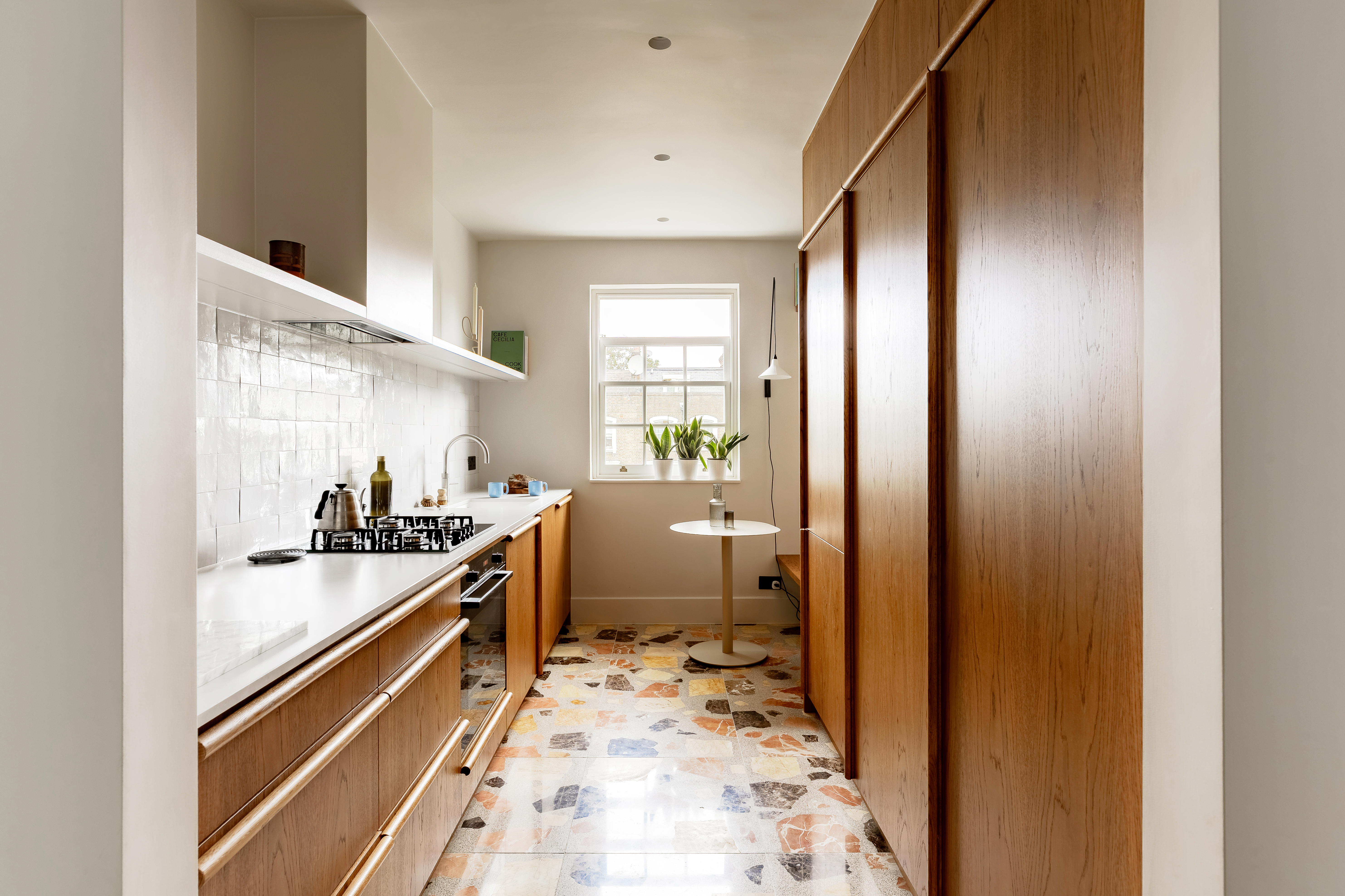 A galley kitchen with terrazzo floor tiles, wooden cabinets and an in-built bench by the window