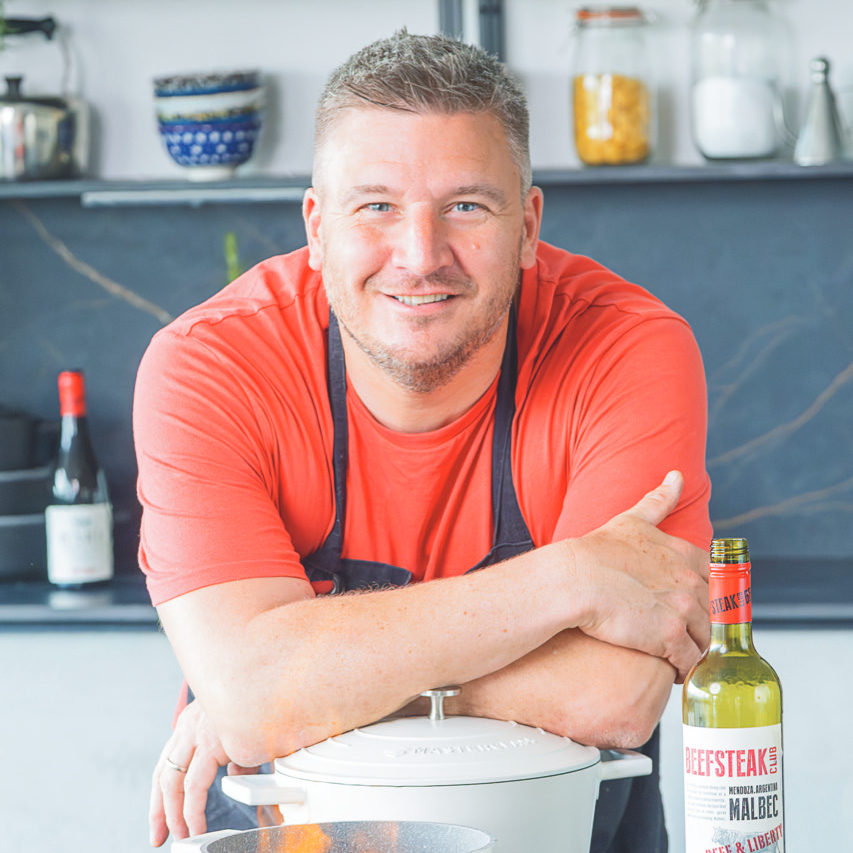 male chef in kitchen wearing red tshirt and navy apron leaning on a cooking pot