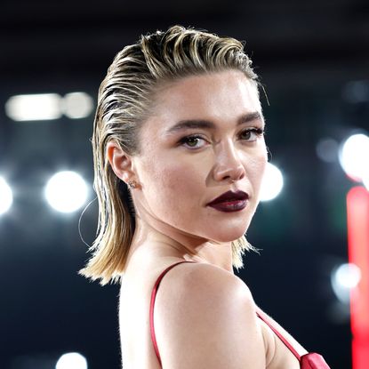 Florence Pugh has slicked back blonde hair with dark roots and wears a dark red lipstick and a red strappy dress