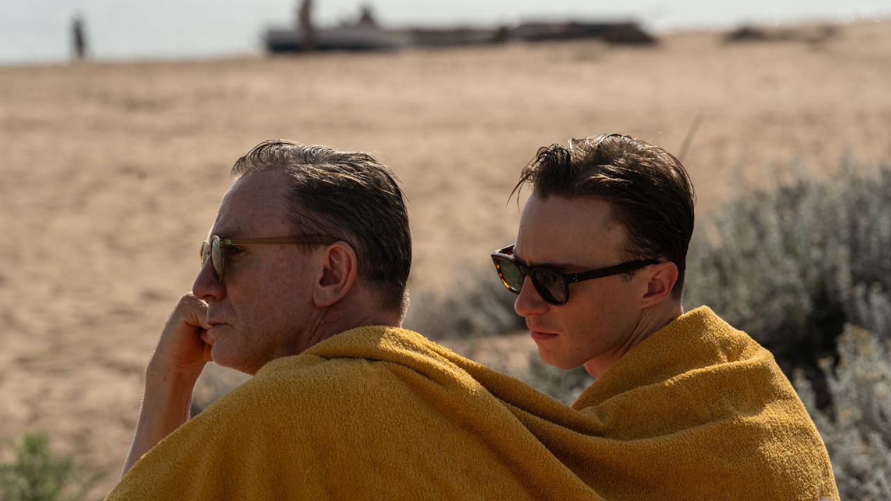 From left to right: Daniel Craig and Drew Starkey sitting on a beach together under a yellow towel.