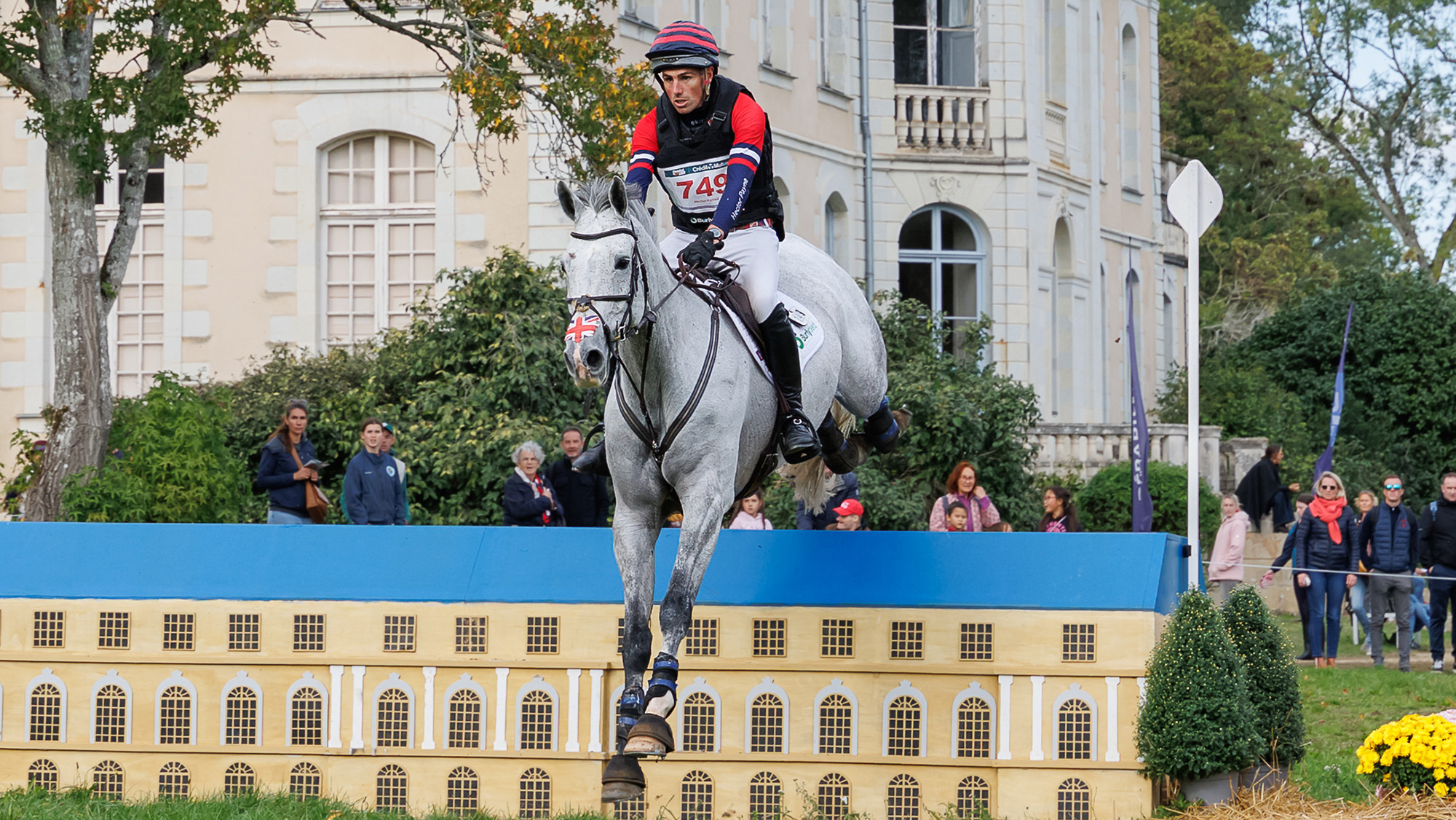 Hector Payne, a rider in red, white and blue striped top and hat, rides grey horse Bueno over a cross-country fence decorated as a house, at Le Lion d&lsquo;Angers 2026
