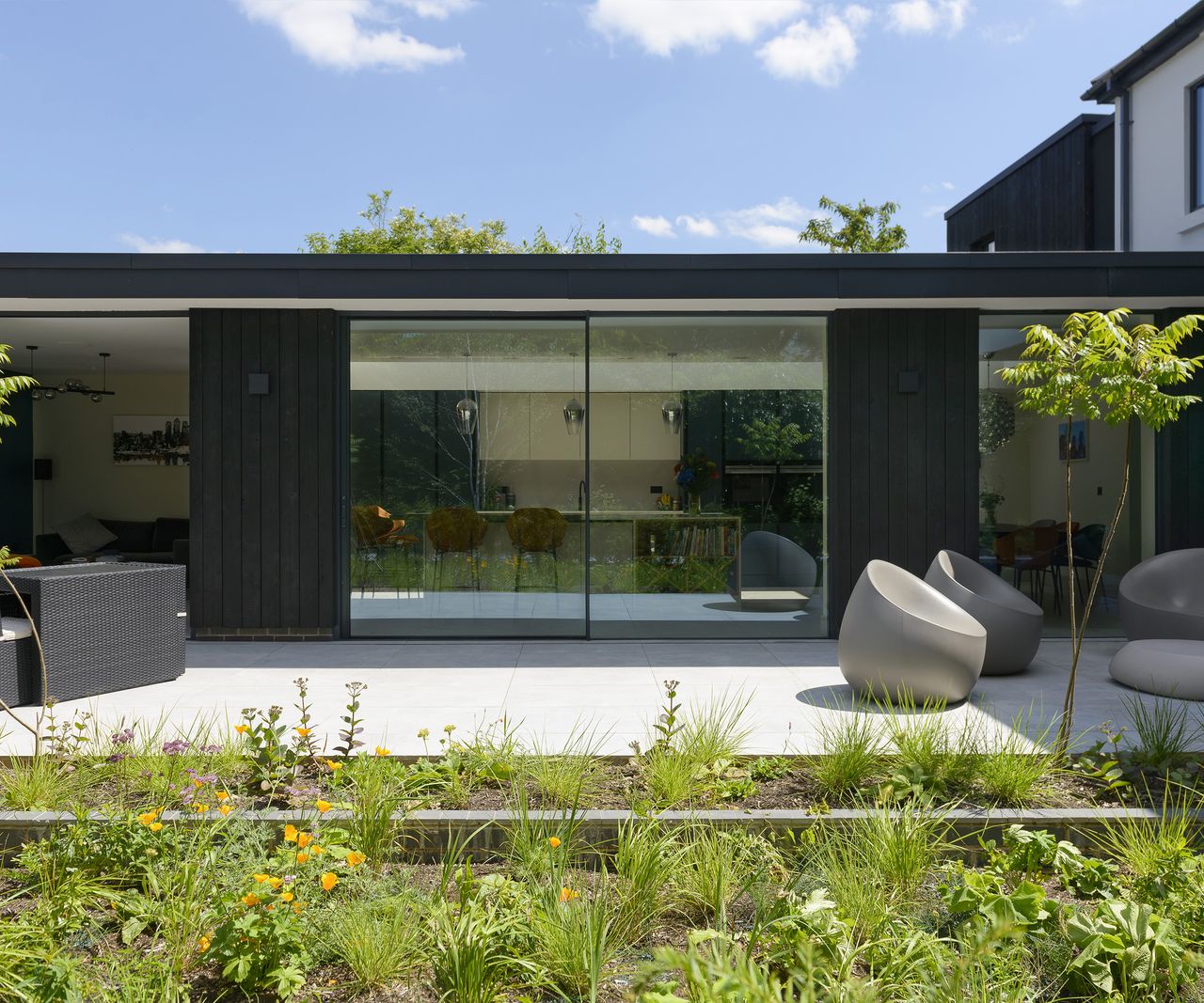 A contemporary extension clad in dark timber and large glazing with a pale grey patio in front with modern garden furniture
