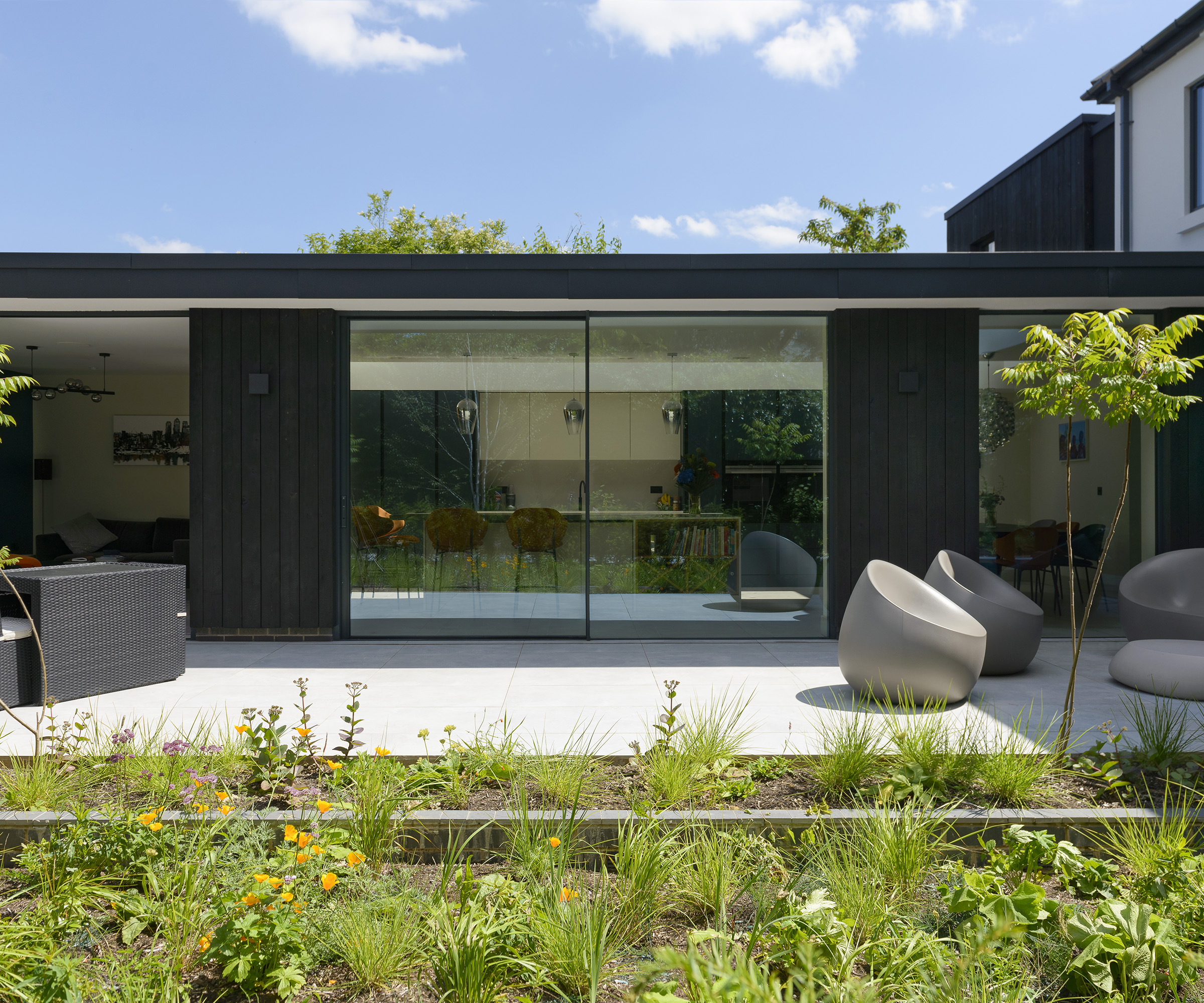 A contemporary extension clad in dark timber and large glazing  with a pale grey patio in front with modern garden furniture