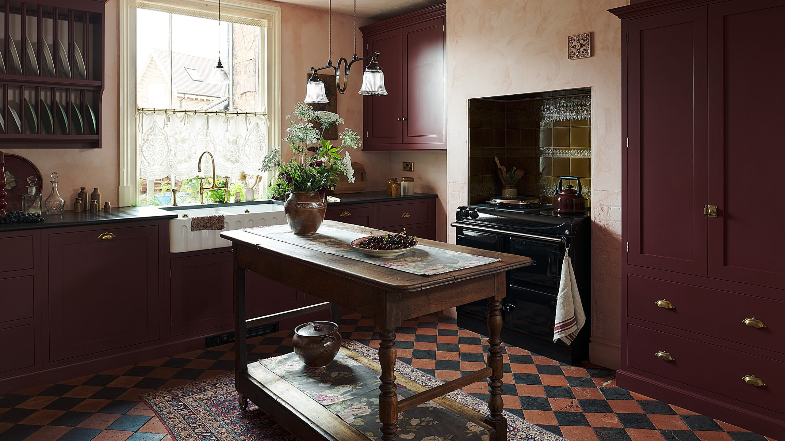 A cosy burgundy kitchen layered with rugs and cafe curtains 