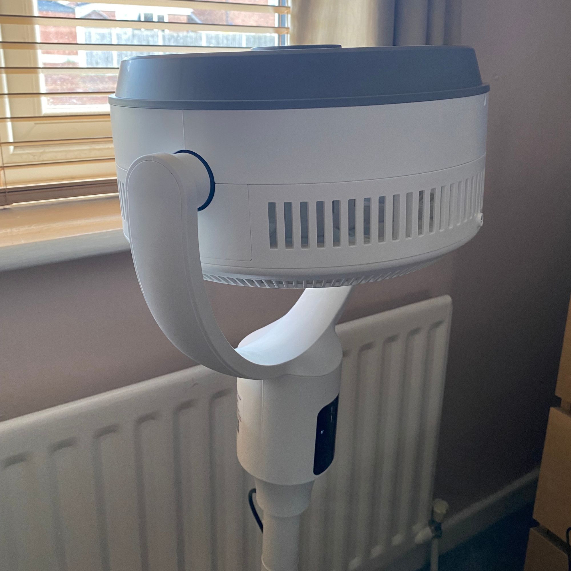 Pro Breeze Airflo 43'' Hybrid Pedestal Fan in the maximum upward tilt position next to a radiator in a plaster pink bedroom with wooden blinds on the window