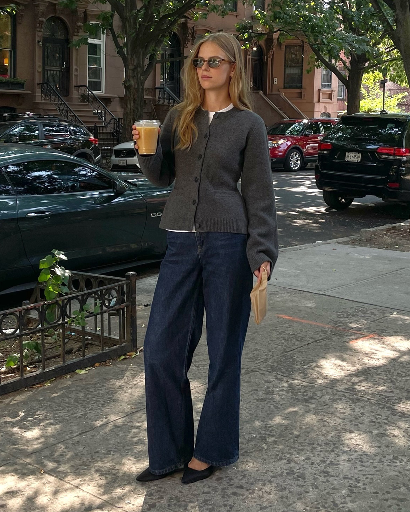 Scandi style influencer and creative Clara Dyrhauge poses on a New York City sidewalk holding a cup of coffee wearing a fitted gray cardigan sweater and relaxed dark-wash jeans