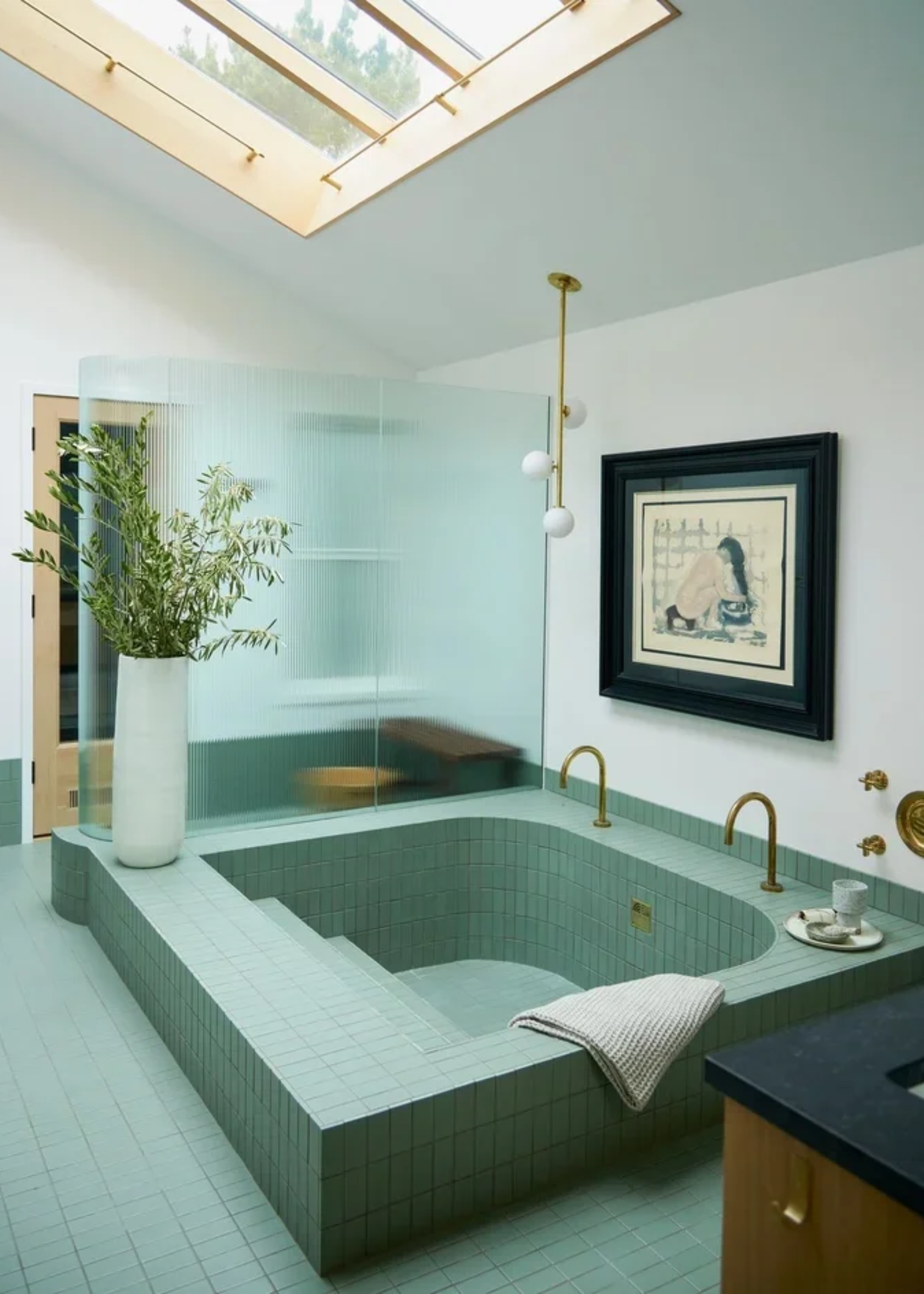 A white bathroom with a skylight and a sea green tiled sunken tub with brass taps by a white vase of stems
