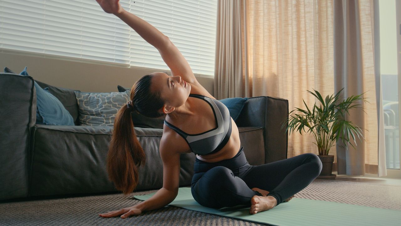 Woman sitting cross-legged on a yoga mat in front of a sofa stretches one arm overhead and to the side. She is wearing athletic leggings and crop top and her brown hair is in a pony tail.