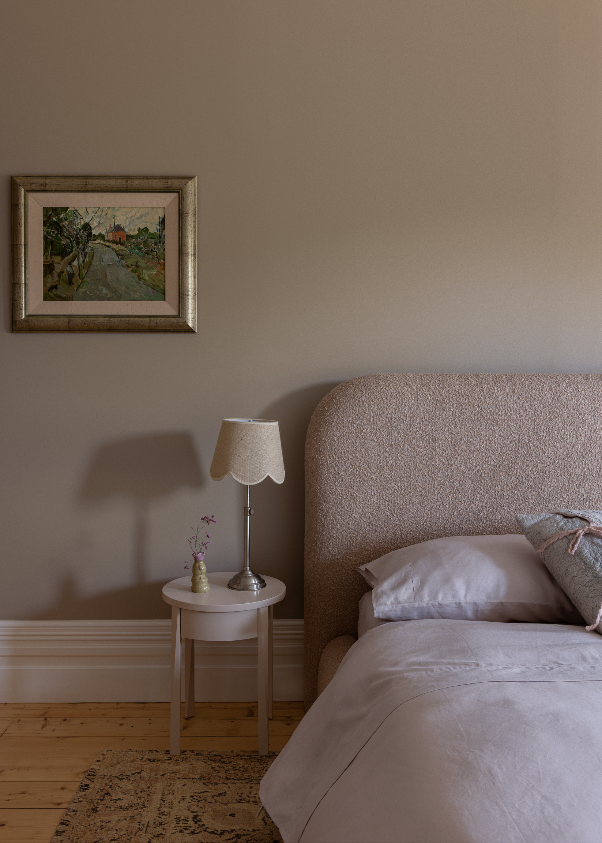 A view of the corner of a bed in a bedroom with a bedside table next to it and a lamp on top of the table with a small flower. There is also artwork hanging on the wall.