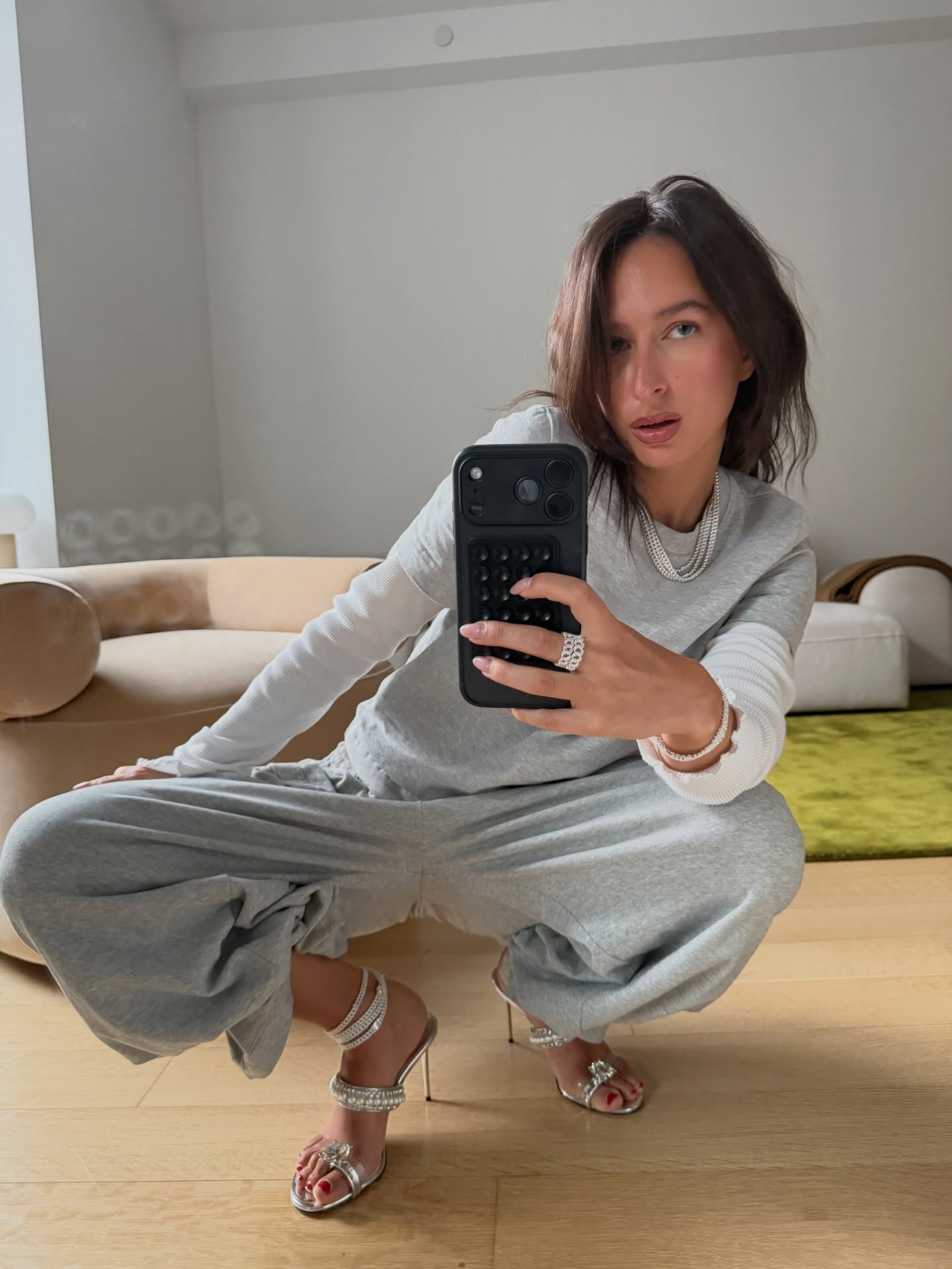 Influencer wearing gray sweatpants, gray t-shirt, white longsleeve, and sparkly silver heels taking a mirror outfit photo.
