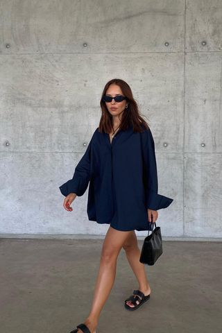 how to make a button-down shirt look more expensive is shown in a photo of a woman standing in a garage wearing black sunglasses, a long necklace, and an oversized navy button-down shirt as a dress that's styled with black slide sandals and a black handbag