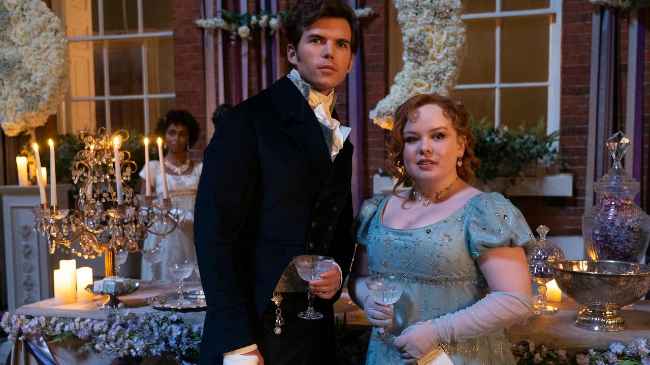 Luke Newton as Colin Bridgerton and Nicola Coughlan as Penelope Featherington, standing by a ball&#039;s drink table with another woman behind them