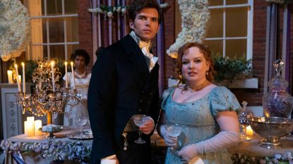 Luke Newton as Colin Bridgerton and Nicola Coughlan as Penelope Featherington, standing by a ball's drink table with another woman behind them