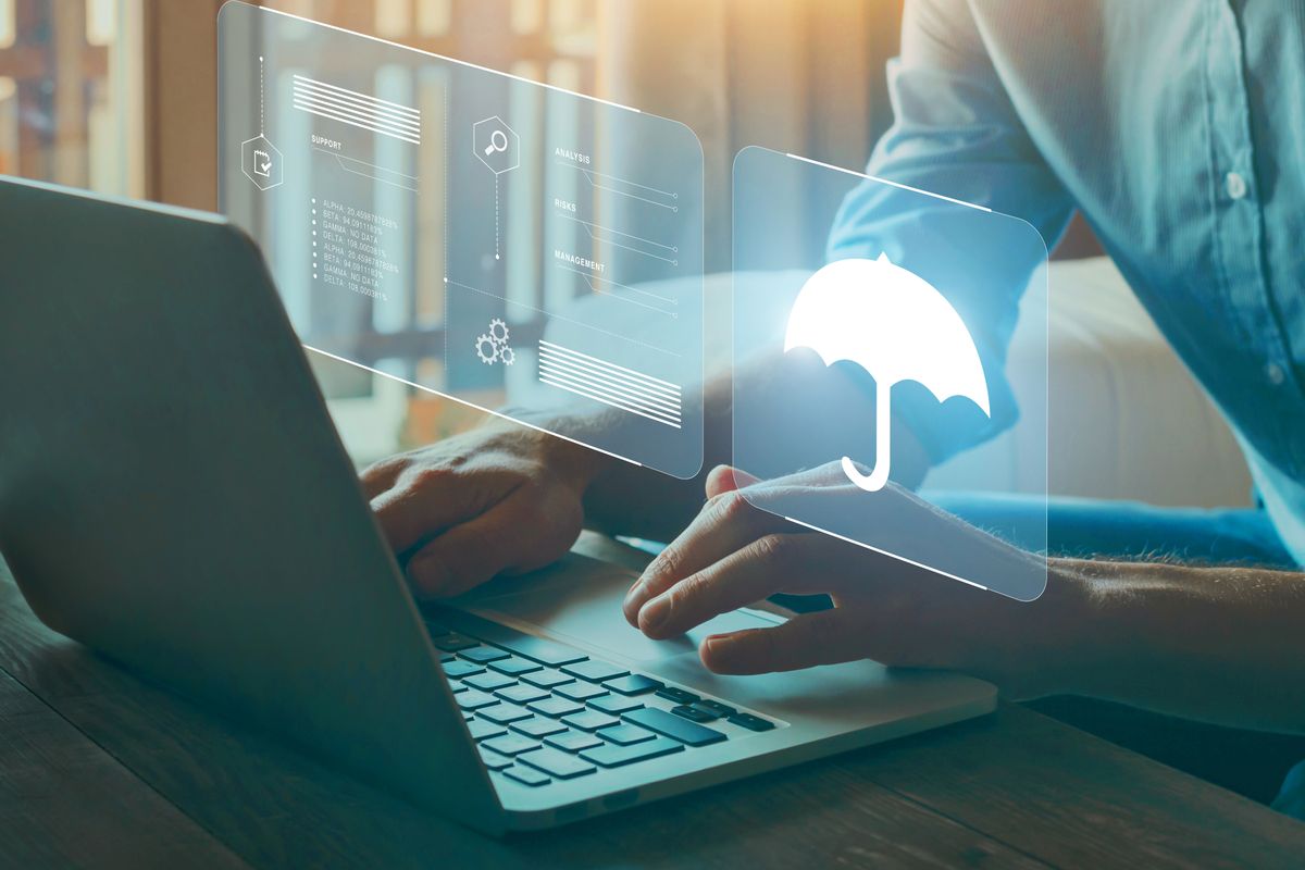 A cyber security insurance and risk management mockup. A person&#039;s hands and torso shown using a laptop, which is seen from behind, as a digital graphic of information and an umbrella icon hover above the keyboard.