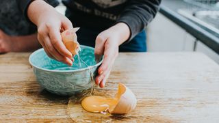 person breaking egg against bowl in kitchen and it spilling onto the side