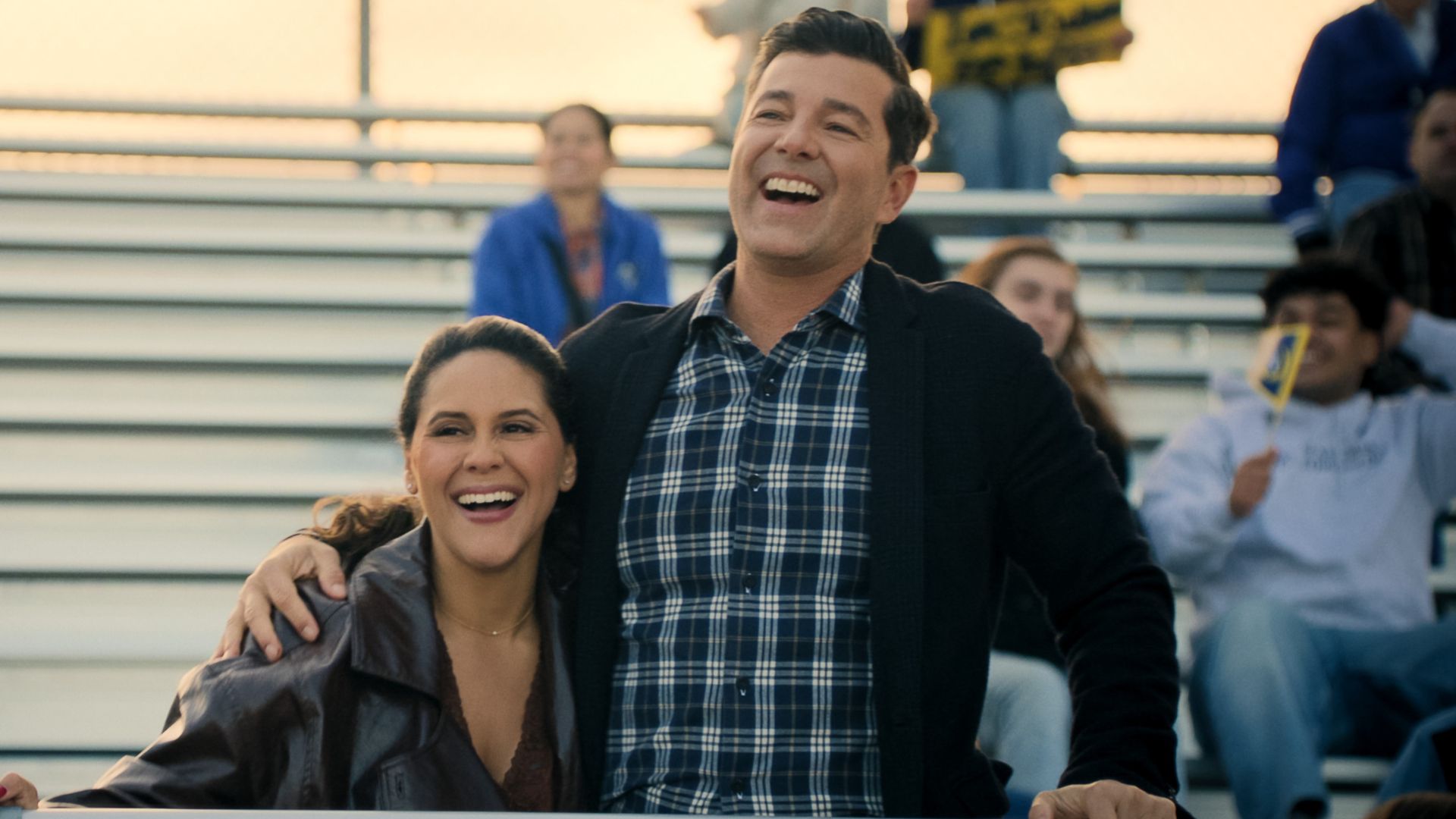 A man and woman embrace in front of bleachers