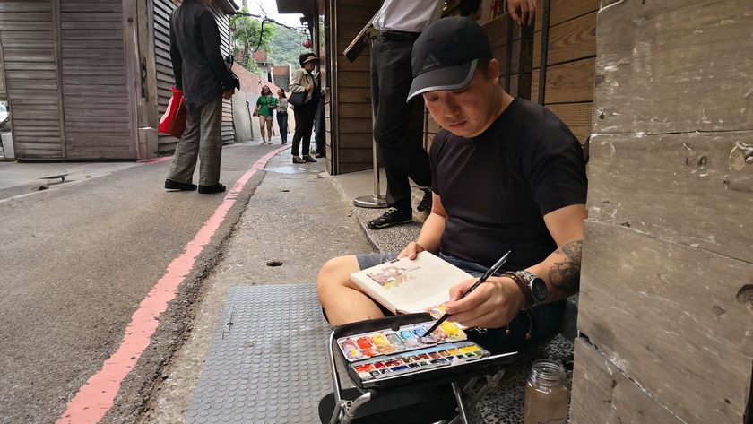 Artist sketchbook tips; a man sits on a stool in a street painting into a sketchbook