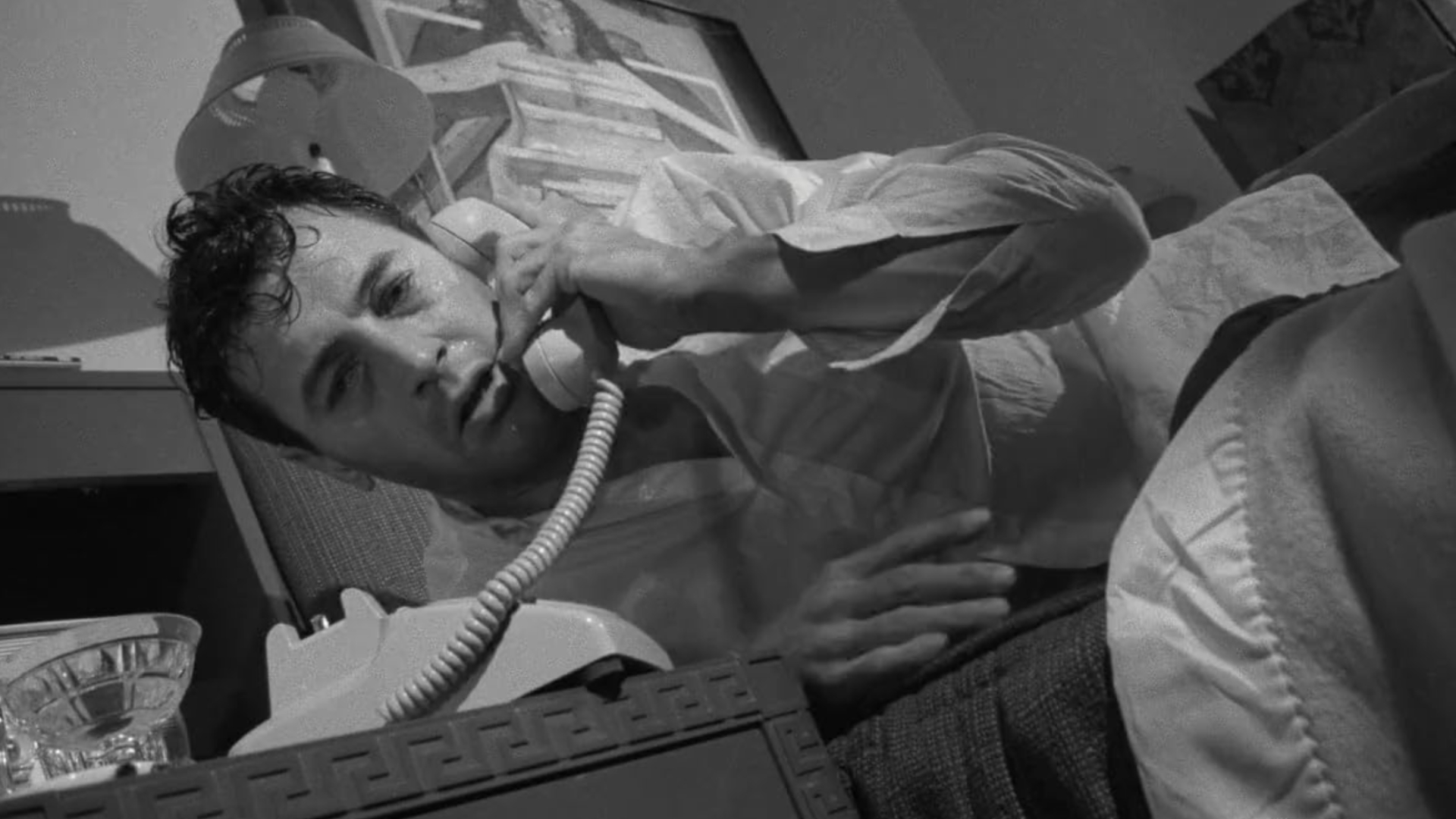 a man wearing a white shirt lies in bed, with a sweaty forehead, answering the phone at his bedside