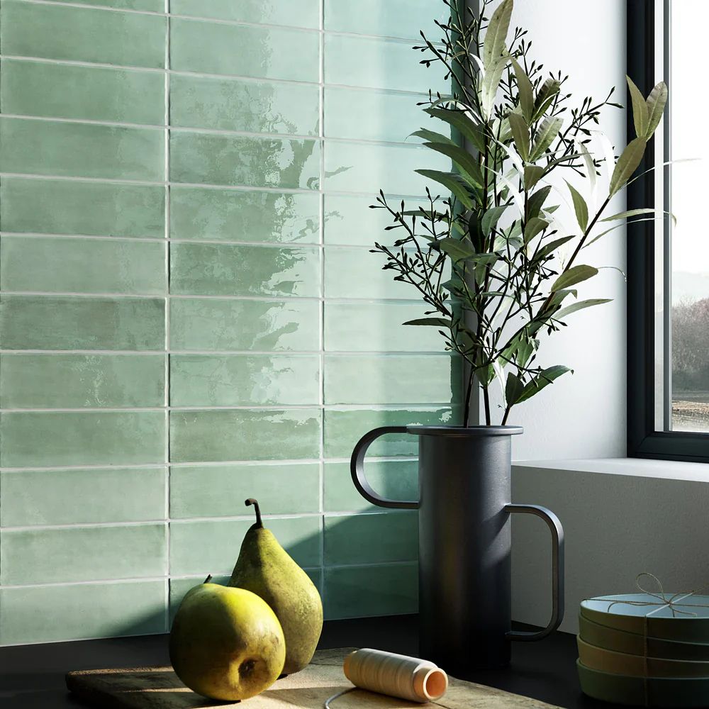 A close-up of glossy sage green rectangular tiles with a handmade, slightly uneven glaze finish laid in a stacked pattern. A black metal jug holding leafy branches, two pears and stacked plates are arranged on a dark worktop beside a black-framed window.