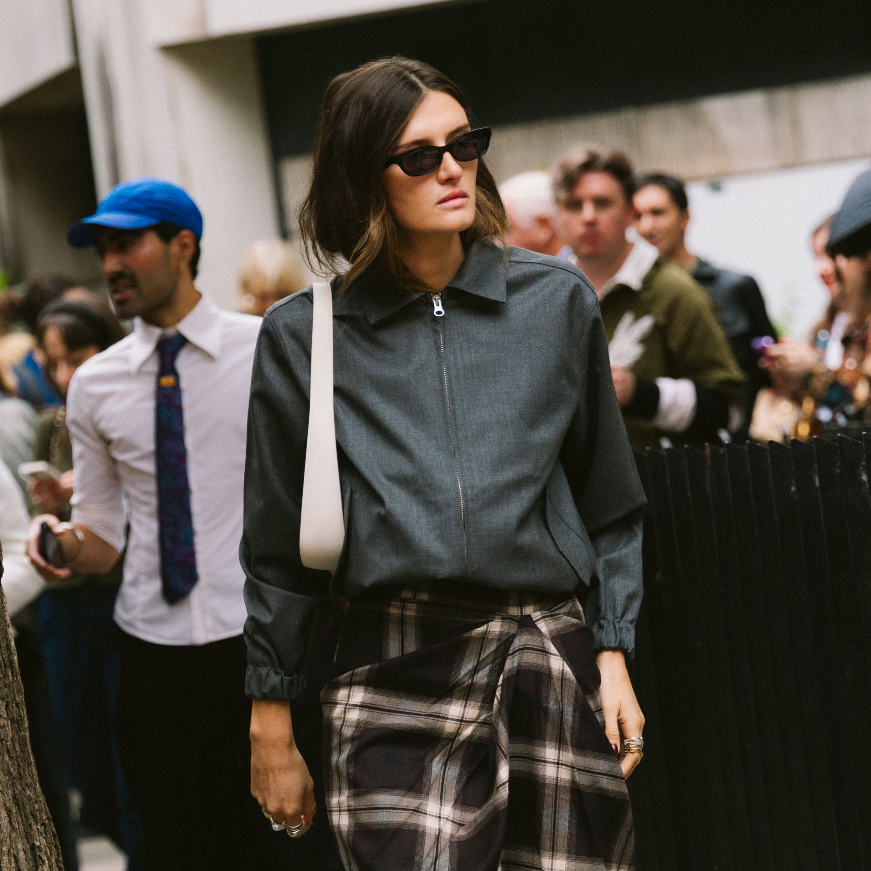 A woman wearing a gray jacket, white bag, and plaid skirt at London Fashion Week
