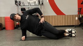 Man demonstrates side plank on his knees. He wears Nike trainers, black pants, black sweatshirt and black cap.